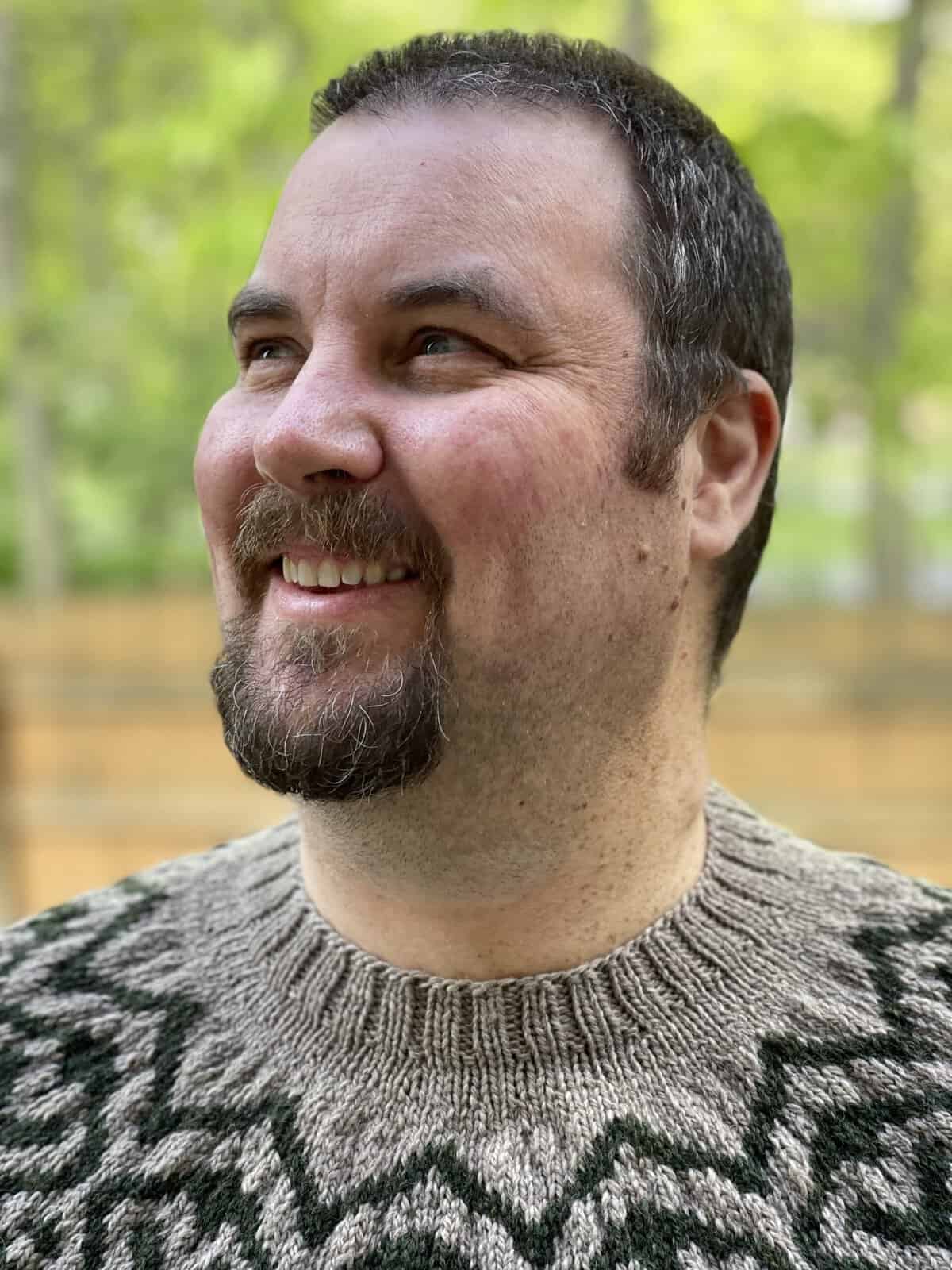 A smiling man in a patterned sweater with trees behind him